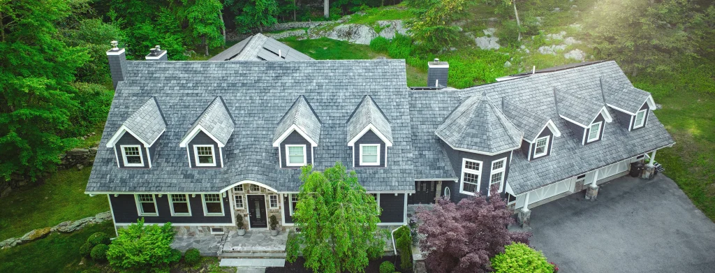 A large two-story house with gray shingles, multiple dormer windows, attached garage, surrounded by trees and greenery.