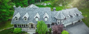A large two-story house with gray shingles, multiple dormer windows, attached garage, surrounded by trees and greenery.
