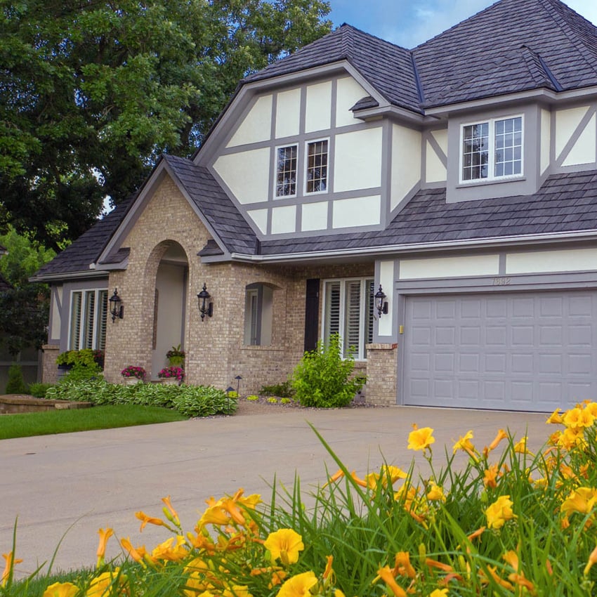 A two-story Tudor-style house with brick and white paneling, a gray garage door, manicured lawn, and yellow flowers in the foreground.
