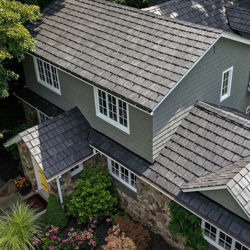A two-story house with a gray shingle roof, gray siding, stone lower walls, white-framed windows, and a yellow front door surrounded by greenery and pink flowers.