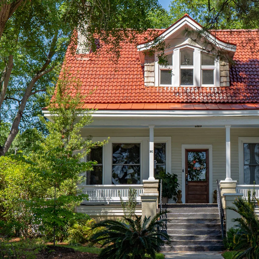 A two-story house with a red tiled roof, white siding, a covered front porch, and stairs leading to a wooden front door, surrounded by trees and shrubs.