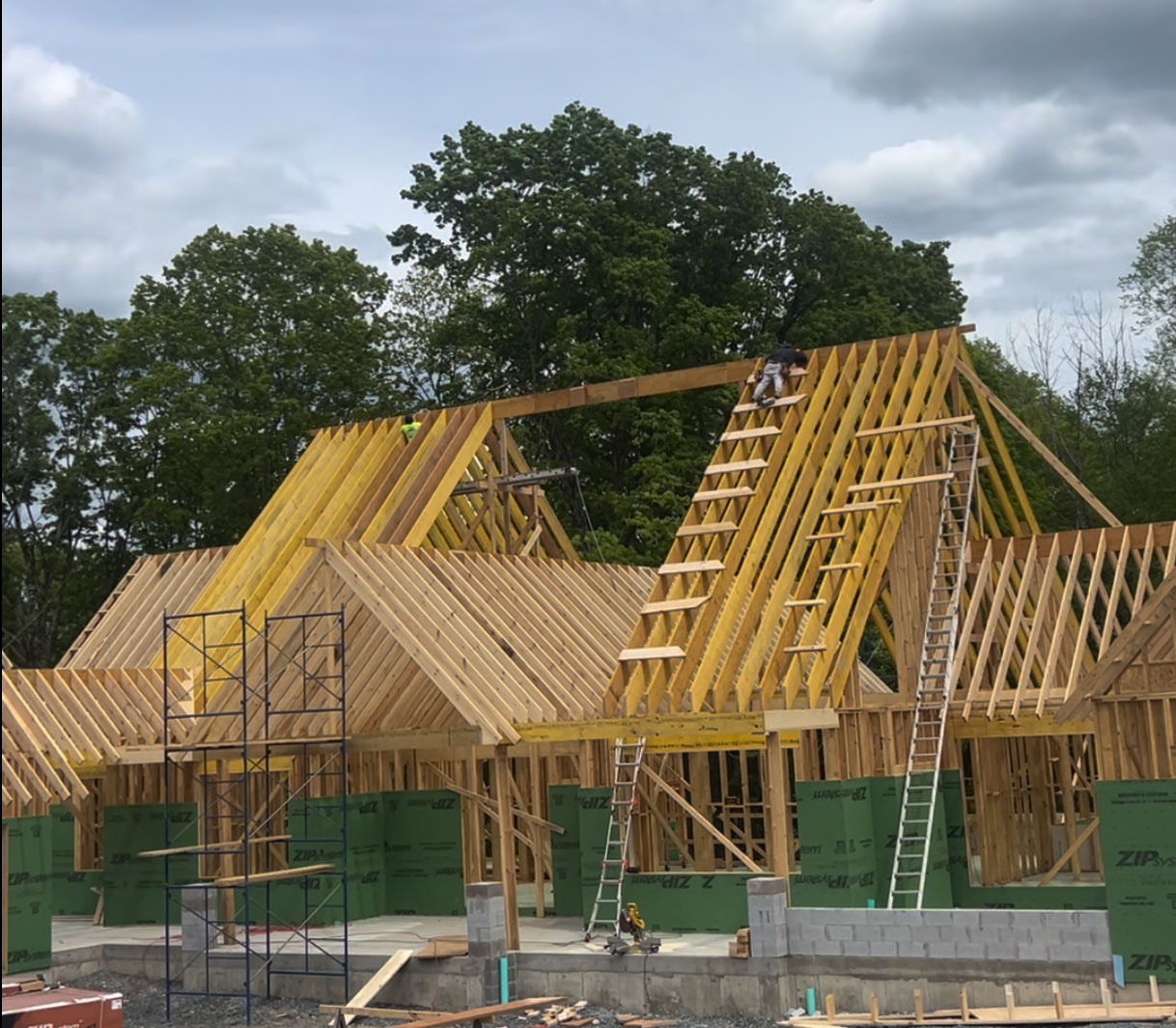 Partially constructed house with exposed wooden framing and roof trusses, ladders, scaffolding, and workers visible against a backdrop of green trees.